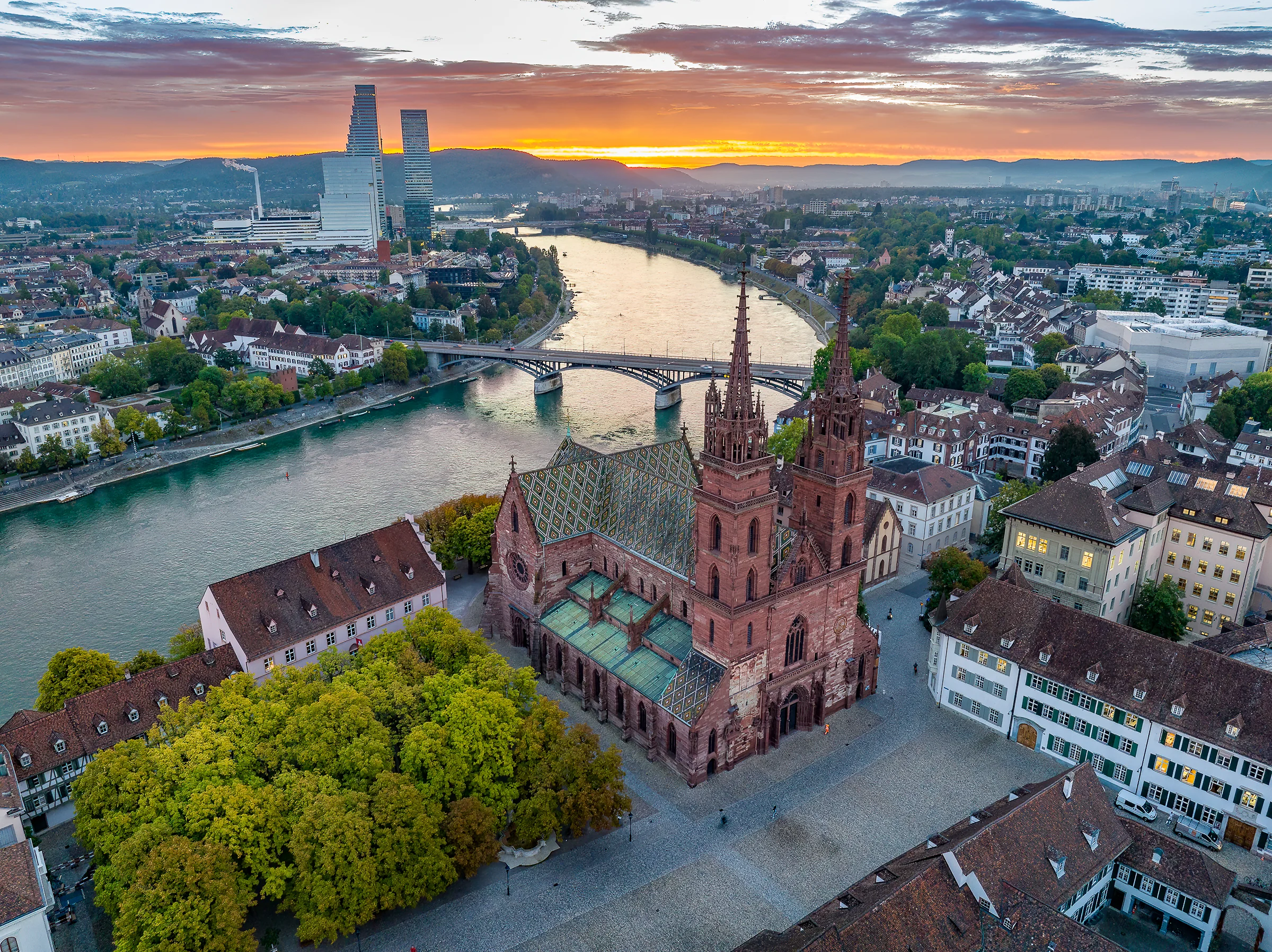 Drohnenaufnahme des Basler Münsters mit rotem Sandsteindach und Spiegelung im Rhein bei goldenem Sonnenuntergang