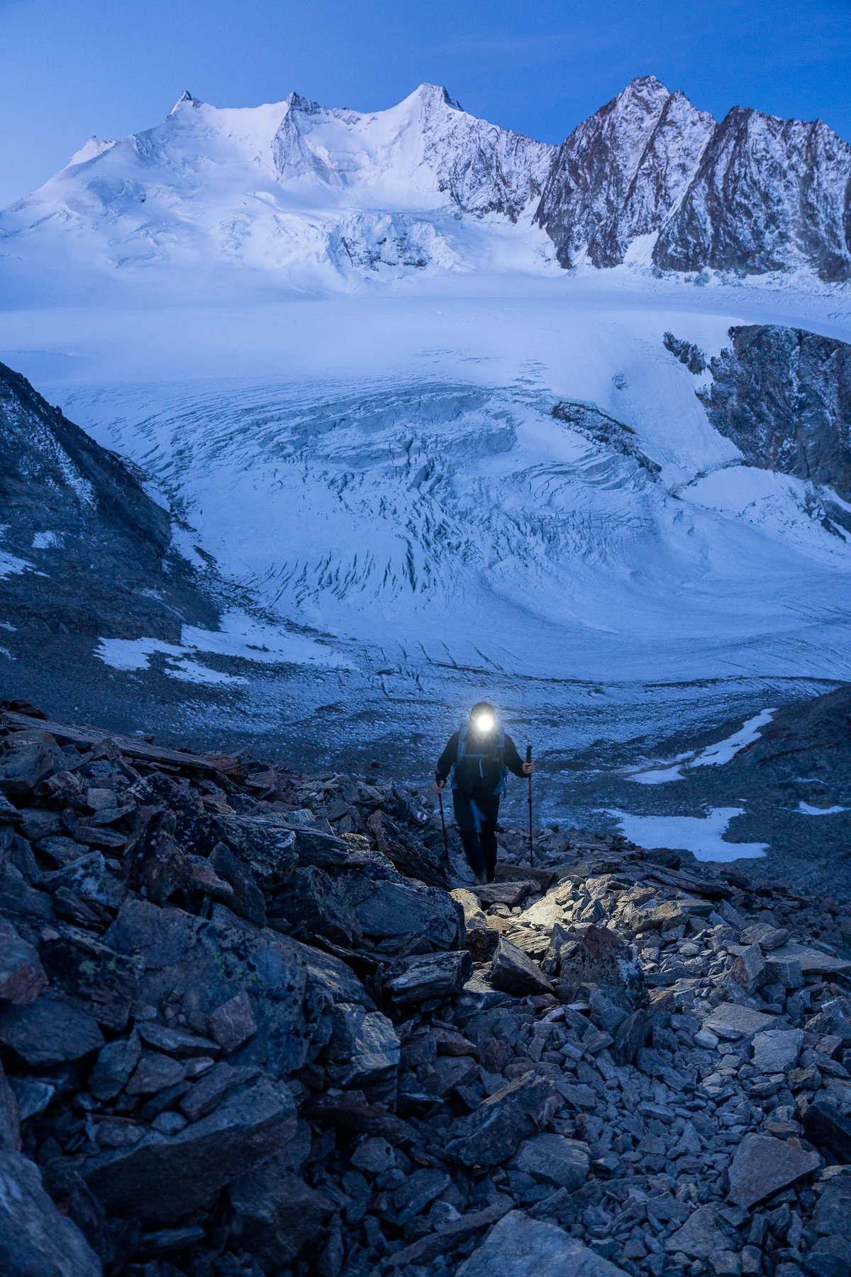 Bergsteiger mit Stirnlampe wandert über schneebedeckten Riedgletscher mit Alpengipfeln im Hintergrund bei Sonnenaufgang
