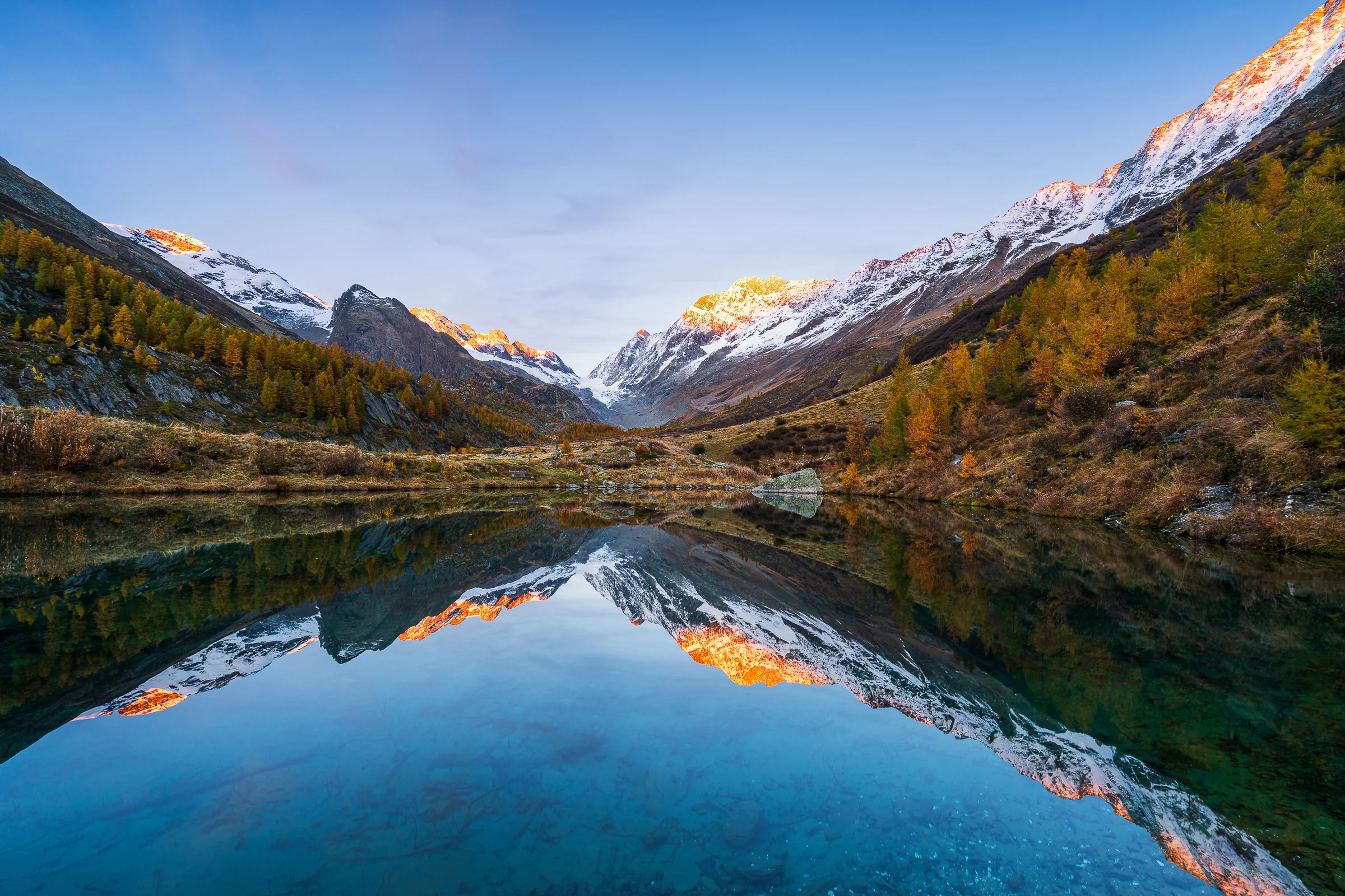 Herbstlicher Bergsee im Lötschental mit spiegelndem Wasser und verschneiten Alpengipfeln bei Sonnenaufgang
