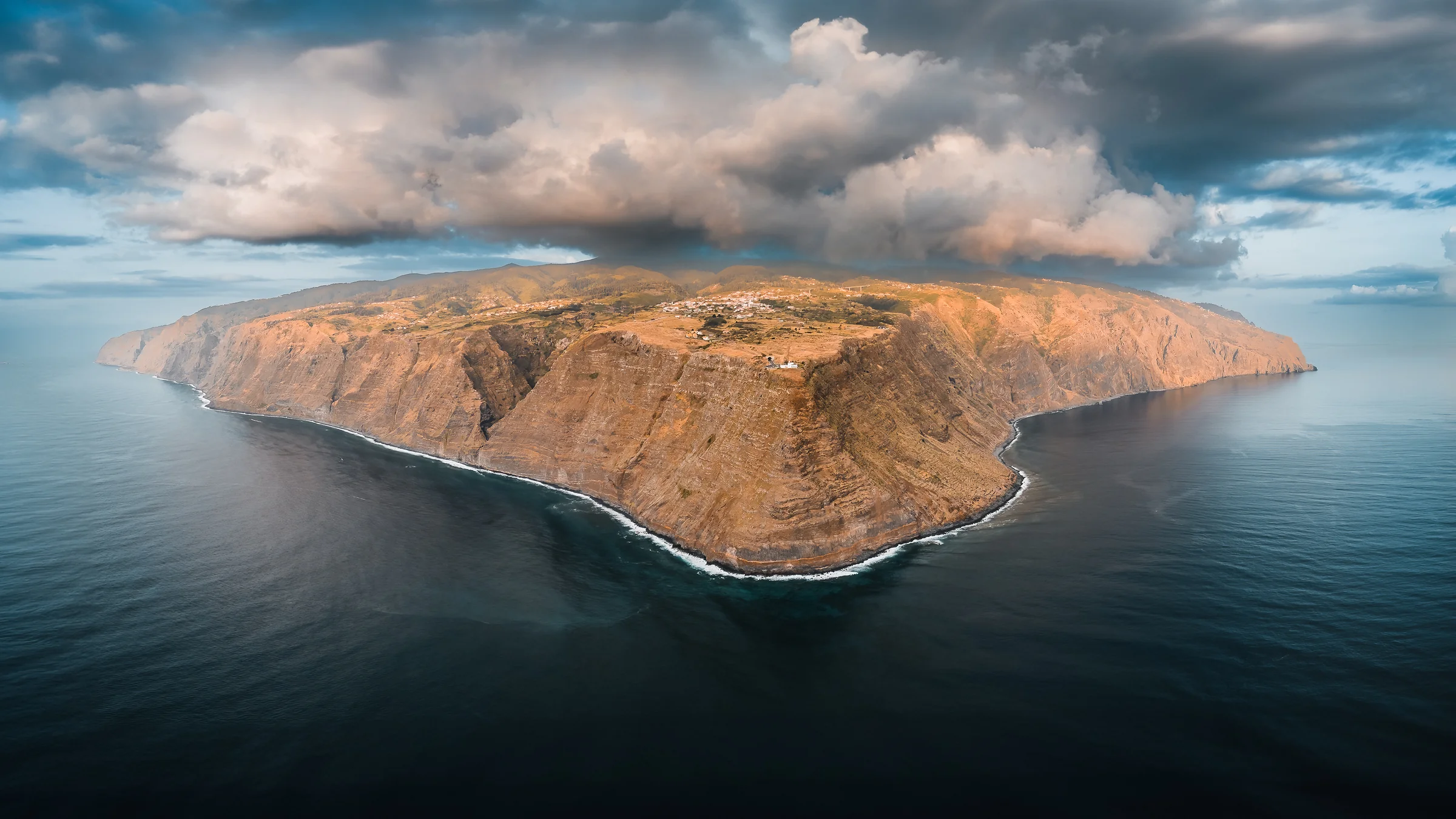 Luftaufnahme der felsigen Madeira-Küste mit steilen Klippen, umgeben von blauem Meer und dramatischen Wolken bei Sonnenuntergang