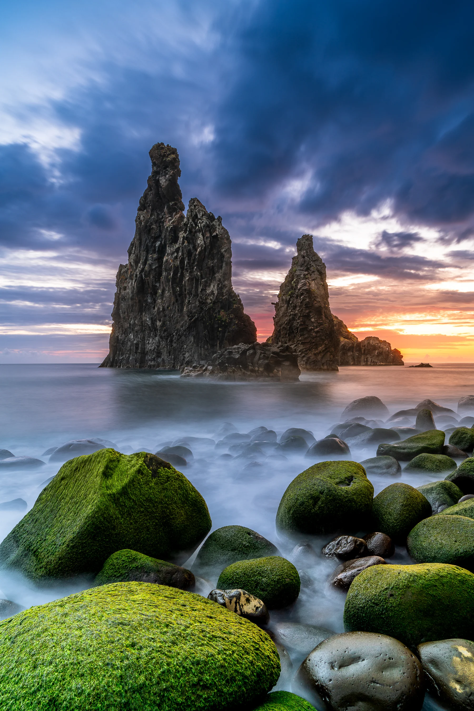 Dramatische Felsnadeln an der Madeira-Küste bei Sonnenaufgang mit moosbedeckten Steinen im Vordergrund und türkisblauem Meer