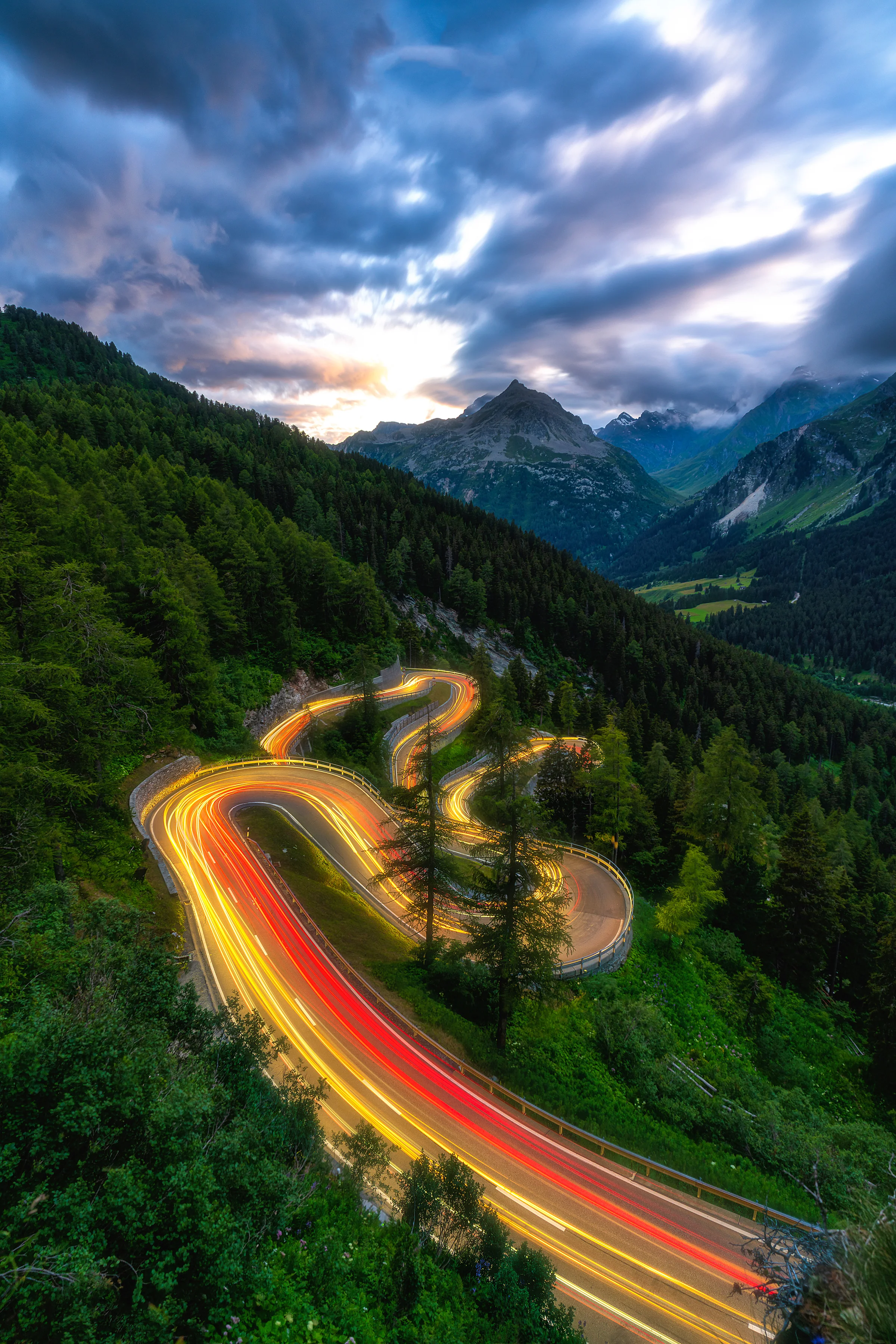 Langzeitbelichtung der gewundenen Maloja Passstrasse mit goldenen und roten Lichtspuren, umgeben von Waldresen und...