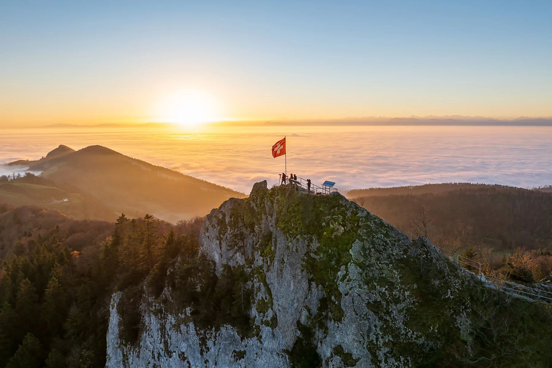 Schweizer Flagge auf dem Felsgipfel der Belchenflue bei Sonnenaufgang über einem Nebelmeer im Baselland
